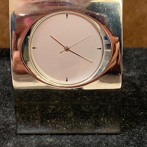May include: A square, silver desk clock with a pale pink face and gold hands. The clock has a minimalist design with a circular face set within a square frame. The clock is a decorative item for a desk or shelf.