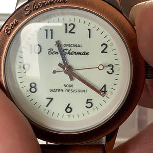 K&ouml;nnte beinhalten: Eine Ben Sherman-Uhr mit kupferfarbenem Geh&auml;use und braunem Lederarmband. Das cremefarbene Zifferblatt hat schwarze Ziffern und Zeiger. Der Text "THE ORIGINAL Ben Sherman" und "50M WATER RESISTANT" sind zu sehen.
