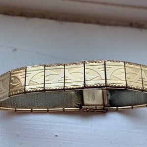 Puede incluir: Un brazalete expandible en tono dorado con un patrón repetido de peces estilizados. El brazalete tiene un cierre con la palabra "SWIZA" grabada en él.