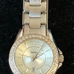 May include: A silver Fossil watch with a white face and crystal accents around the bezel. The watch has a silver metal band.