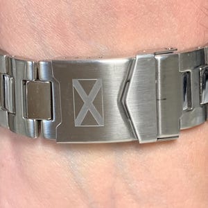 Puede incluir: Una correa de reloj de metal plateado con un dise&ntilde;o rectangular. El cierre presenta un s&iacute;mbolo 'X' grabado. La correa est&aacute; sujeta a una mu&ntilde;eca, mostrando el acabado met&aacute;lico y el dise&ntilde;o intrincado del reloj. Un accesorio elegante.