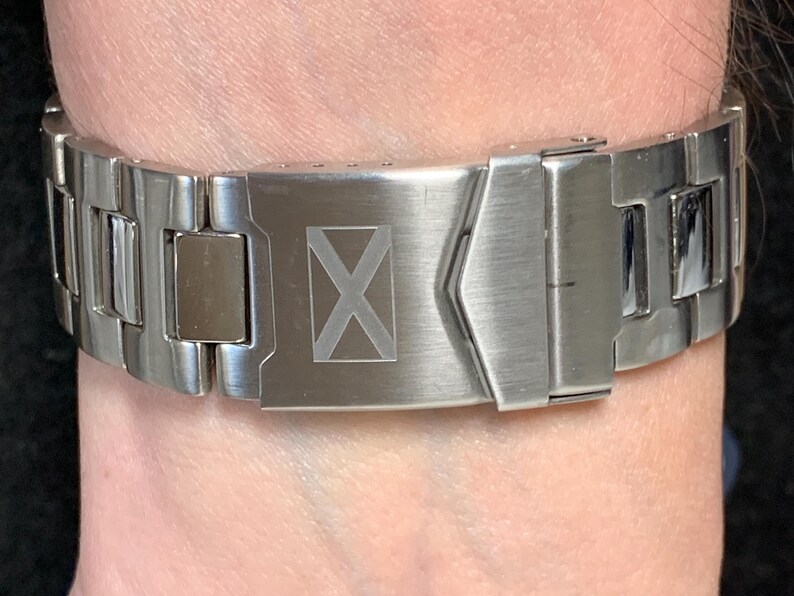 Puede incluir: Una correa de reloj de metal plateado con un dise&ntilde;o rectangular. El cierre presenta un s&iacute;mbolo 'X' grabado. La correa est&aacute; sujeta a una mu&ntilde;eca, mostrando el acabado met&aacute;lico y el dise&ntilde;o intrincado del reloj. Un accesorio elegante.