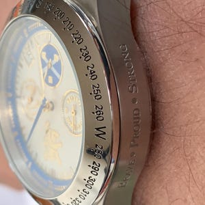 Puede incluir: Primer plano de un reloj de pulsera plateado con esfera blanca y detalles azules. El reloj tiene un bisel tipo br&uacute;jula con marcas direccionales y las palabras "Brave Proud Strong" grabadas en el lateral.