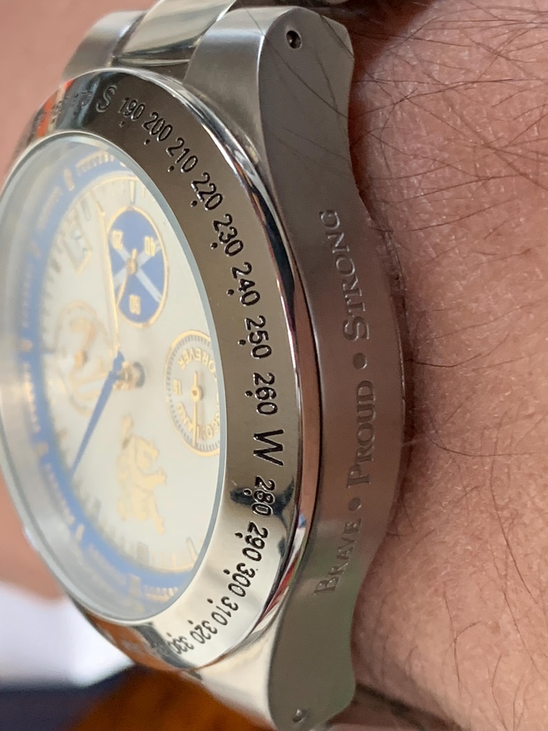 Puede incluir: Primer plano de un reloj de pulsera plateado con esfera blanca y detalles azules. El reloj tiene un bisel tipo br&uacute;jula con marcas direccionales y las palabras "Brave Proud Strong" grabadas en el lateral.
