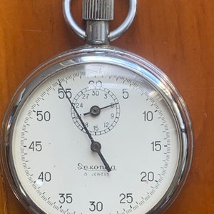Könnte beinhalten: Eine silberne Taschenuhr mit weißem Zifferblatt und schwarzen Zeigern. Die Uhr hat einen schwarzen Sekundenzeiger und ein kleines Zifferblatt mit den Zahlen 1 bis 12. Das Zifferblatt der Uhr hat auch die Zahlen 5, 10, 15, 20, 25, 30, 35, 40, 45, 50, 55 und 60. Das Zifferblatt der Uhr hat auch den Text "Sekonda 15 Jewels" und "Made in USSR".