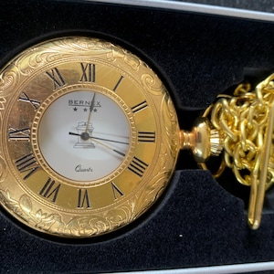 Puede incluir: Reloj de bolsillo dorado con números romanos y detalles ornamentados. La esfera blanca muestra la marca "BERNEX" y agujas negras. Incluye cadena y cierre dorados, presentado en una caja negra. Movimiento de cuarzo.