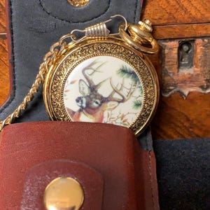 Op de afbeelding: Een goudkleurig zakhorloge met een illustratie van een hert op een crèmekleurige wijzerplaat. Het heeft een sierlijke rand en een gouden ketting. Het horloge ligt in een bruin lederen etui met een gouden knoop en een zwart interieur.