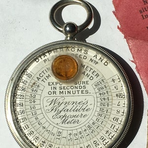 Op de afbeelding: Een vintage Wynne's Infallible Belichtingsmeter, cirkelvormig met een zilverkleurige metalen behuizing. De wijzerplaat toont diafragma- en tijdsinstellingen in zwart. Het heeft een amberkleurige lens en een ring om op te hangen. Diameter ongeveer 6 cm.
