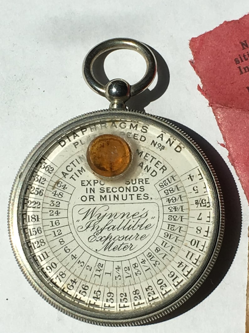 Op de afbeelding: Een vintage Wynne's Infallible Belichtingsmeter, cirkelvormig met een zilverkleurige metalen behuizing. De wijzerplaat toont diafragma- en tijdsinstellingen in zwart. Het heeft een amberkleurige lens en een ring om op te hangen. Diameter ongeveer 6 cm.