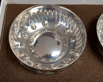 May include: A set of two silver bowls with a decorative embossed design. The bowls have a smooth, reflective surface and a raised rim.