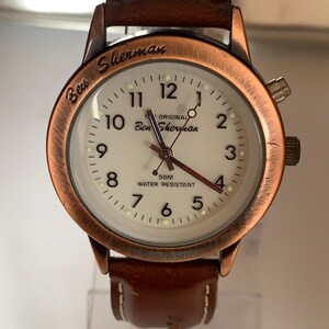 K&ouml;nnte beinhalten: Eine Ben Sherman Armbanduhr mit wei&szlig;em Zifferblatt, kupferfarbener L&uuml;nette und braunem Lederarmband. Das Zifferblatt zeigt den Markennamen und "50M Water Resistant". Die Uhr hat ein klassisches Design.