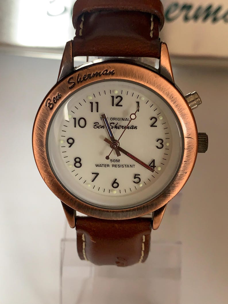 K&ouml;nnte beinhalten: Eine Ben Sherman Armbanduhr mit wei&szlig;em Zifferblatt, kupferfarbener L&uuml;nette und braunem Lederarmband. Das Zifferblatt zeigt den Markennamen und "50M Water Resistant". Die Uhr hat ein klassisches Design.