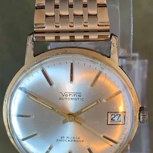 Puede incluir: Un reloj de pulsera dorado con una esfera blanca, agujas negras y una ventana de fecha que muestra el número 27. La esfera del reloj también presenta el texto "Vehns Automatic" y "25 Rubis Shockproof".