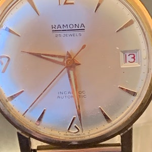 May include: Close-up of a vintage Ramona wristwatch. The watch features a gold-toned case and hands, with a silver-colored face. The watch face displays the text "RAMONA 25 JEWELS", and the date is set to 13. The watch has a metal band.