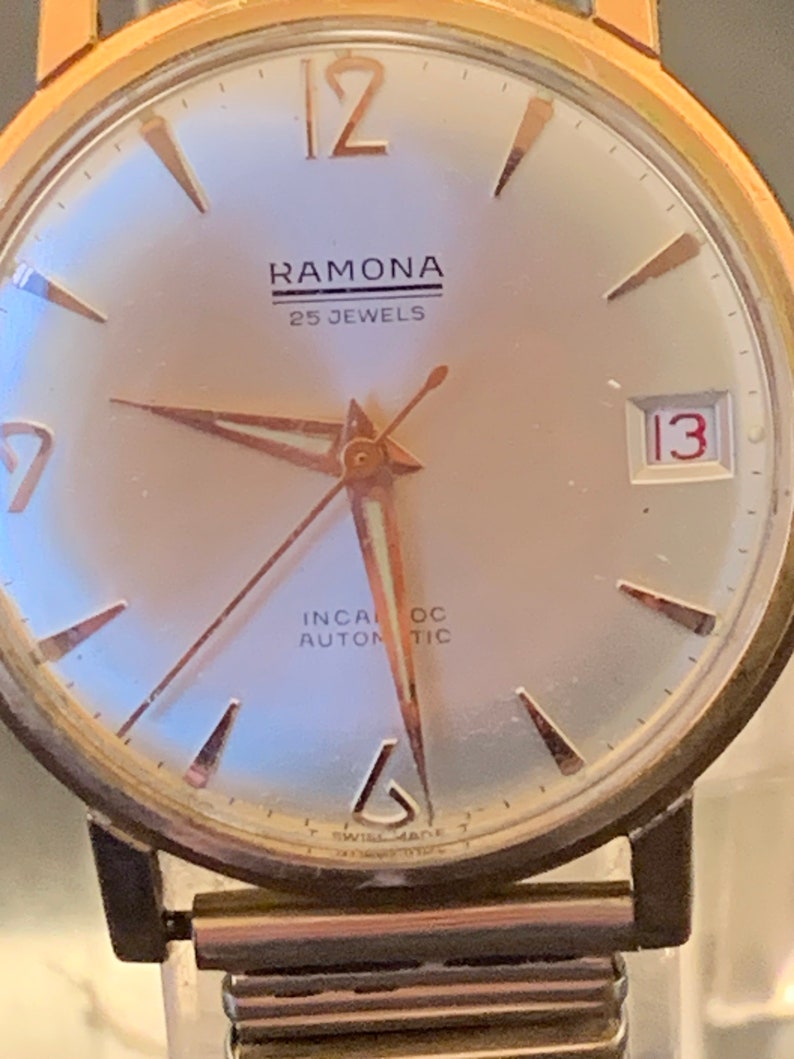 May include: Close-up of a vintage Ramona wristwatch. The watch features a gold-toned case and hands, with a silver-colored face. The watch face displays the text "RAMONA 25 JEWELS", and the date is set to 13. The watch has a metal band.