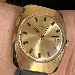 May include: A gold-toned Sekonda wristwatch with a square face and black hands. The watch face is gold with black markings and the text "30 jewels automatic".