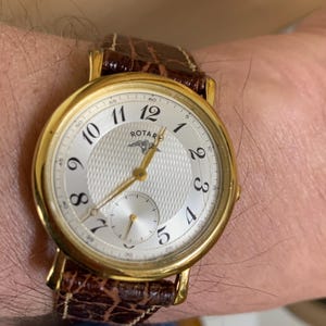 Peut inclure: Une montre-bracelet dorée avec un cadran blanc et un bracelet en cuir marron. Le cadran affiche des chiffres noirs, un petit cadran des secondes et la marque "ROTARD". Le bracelet présente un motif texturé en forme de crocodile.