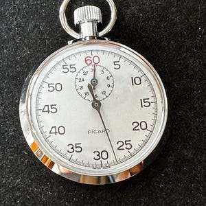 Peut inclure: Un chronomètre argenté avec un cadran blanc, affichant des chiffres noirs et des accents rouges à la marque des 60 secondes. Il comporte un petit cadran secondaire et une couronne de remontoir. La marque "PICARD" est imprimée.