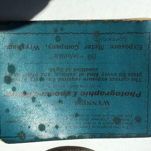Op de afbeelding: Een blauwe vintage fotografische belichtingsmeter met tekst. De tekst bevat "WYNNE'S Photographic Exposure Meter" en "Exposure Meter Company, Wrexham". Het is ontworpen om de juiste belichting te bepalen.