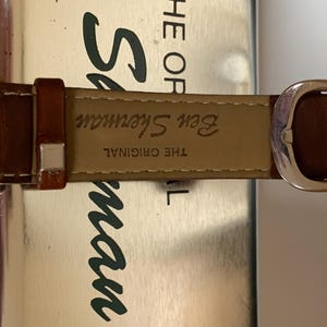 K&ouml;nnte beinhalten: Nahaufnahme eines braunen Leder-Uhrenarmbands mit silberner Schnalle. Auf dem Armband steht "Ben Sherman THE ORIGINAL". Das Zifferblatt ist links teilweise sichtbar. Der Hintergrund ist eine metallische Oberfl&auml;che.