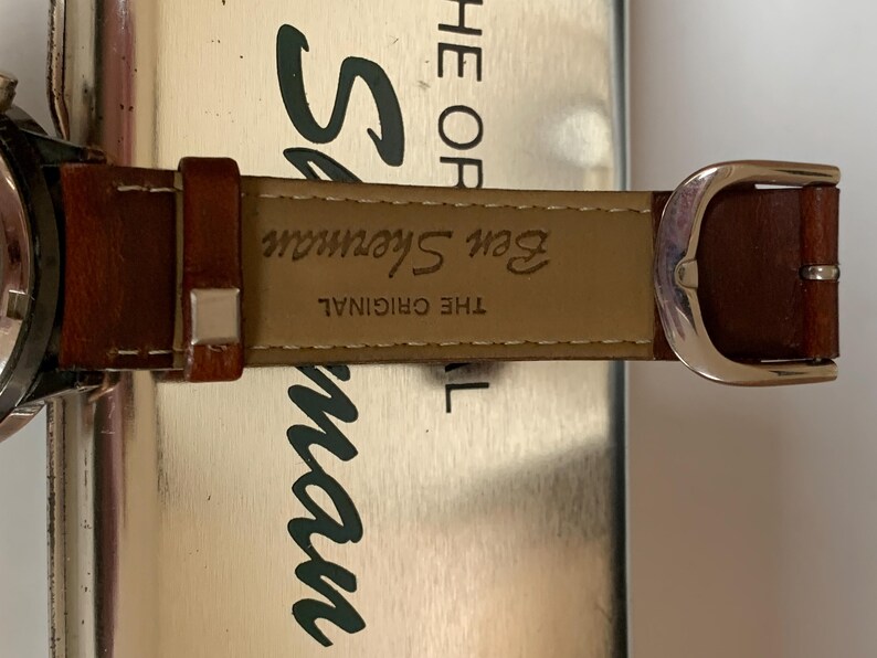 K&ouml;nnte beinhalten: Nahaufnahme eines braunen Leder-Uhrenarmbands mit silberner Schnalle. Auf dem Armband steht "Ben Sherman THE ORIGINAL". Das Zifferblatt ist links teilweise sichtbar. Der Hintergrund ist eine metallische Oberfl&auml;che.