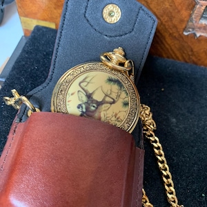 May include: A gold-toned pocket watch with a deer illustration on the face, nestled in a brown leather case with a black interior. A gold chain is attached. The watch is a classic accessory.