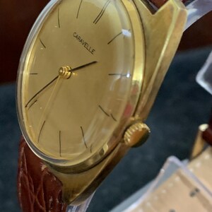 May include: A gold-toned wristwatch with a brown leather band. The watch face is round and has a gold-toned bezel. The watch face has black Roman numeral hour markers and black hands. The watch face also has the word "Caravelle" printed in black.