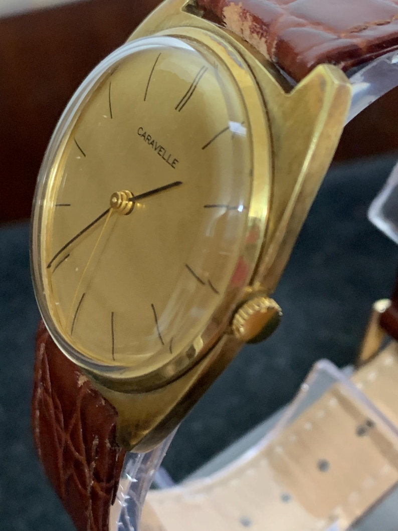 May include: A gold-toned wristwatch with a brown leather band. The watch face is round and has a gold-toned bezel. The watch face has black Roman numeral hour markers and black hands. The watch face also has the word "Caravelle" printed in black.