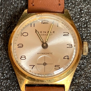Peut inclure: Une montre-bracelet vintage dorée avec un cadran clair et un bracelet en cuir marron. Le cadran affiche la marque "RENZLE ARKANT" et le mot "ANTIMAGNETIC". La montre a un cadran rond avec des chiffres arabes et une trotteuse.