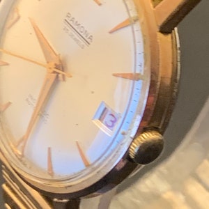 May include: Close-up of a vintage Ramona wristwatch. The watch features a gold-toned case and hands, with a cream-colored face. The watch face displays the number 13 in a small window. The watch band is a gold-toned metal.
