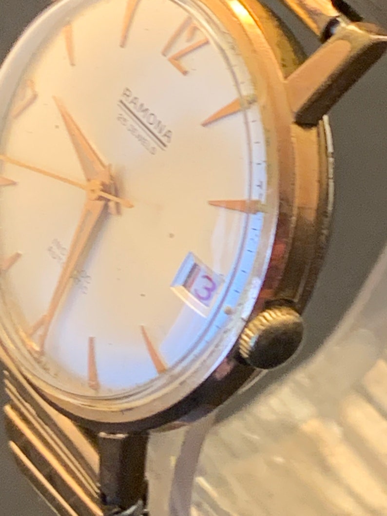May include: Close-up of a vintage Ramona wristwatch. The watch features a gold-toned case and hands, with a cream-colored face. The watch face displays the number 13 in a small window. The watch band is a gold-toned metal.