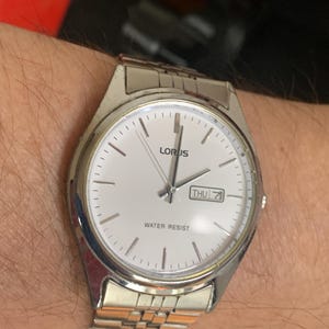 Puede incluir: Un reloj de pulsera Lorus plateado con una esfera blanca, agujas plateadas y una correa plateada. La esfera del reloj tiene una pantalla del día de la semana que dice "THU" y las palabras "WATER RESIST".