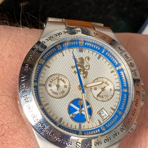 Puede incluir: Reloj de pulsera plateado con esfera blanca, detalles azules y correa de acero inoxidable. El reloj tiene subesferas, visualizaci&oacute;n de fecha y la inscripci&oacute;n "SCOTLAND FOREVER". Tiene un emblema de le&oacute;n dorado.