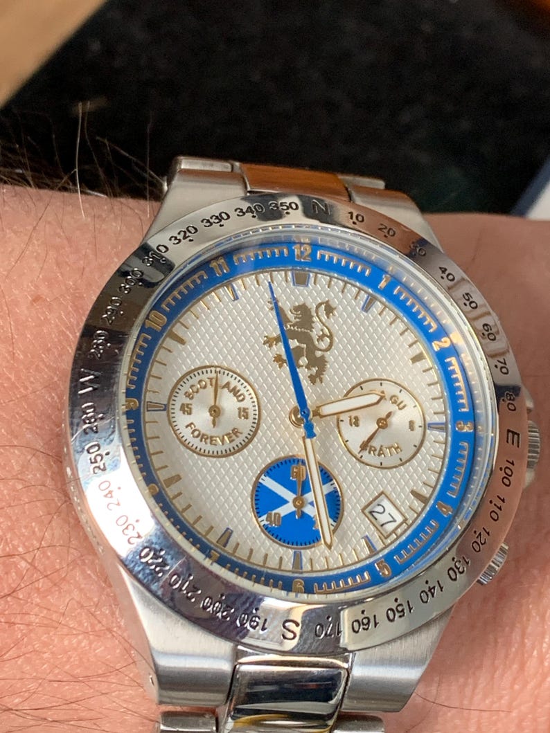Puede incluir: Reloj de pulsera plateado con esfera blanca, detalles azules y correa de acero inoxidable. El reloj tiene subesferas, visualizaci&oacute;n de fecha y la inscripci&oacute;n "SCOTLAND FOREVER". Tiene un emblema de le&oacute;n dorado.