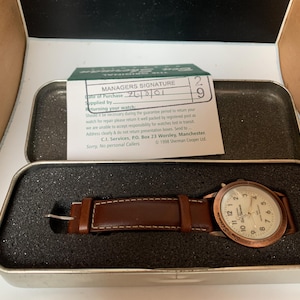 K&ouml;nnte beinhalten: Eine Vintage-Armbanduhr mit braunem Lederarmband und kupferfarbenem Geh&auml;use, pr&auml;sentiert in einer Metalldose. Das Zifferblatt der Uhr ist wei&szlig; mit schwarzen Ziffern. Eine kleine Karte mit Text ist sichtbar.