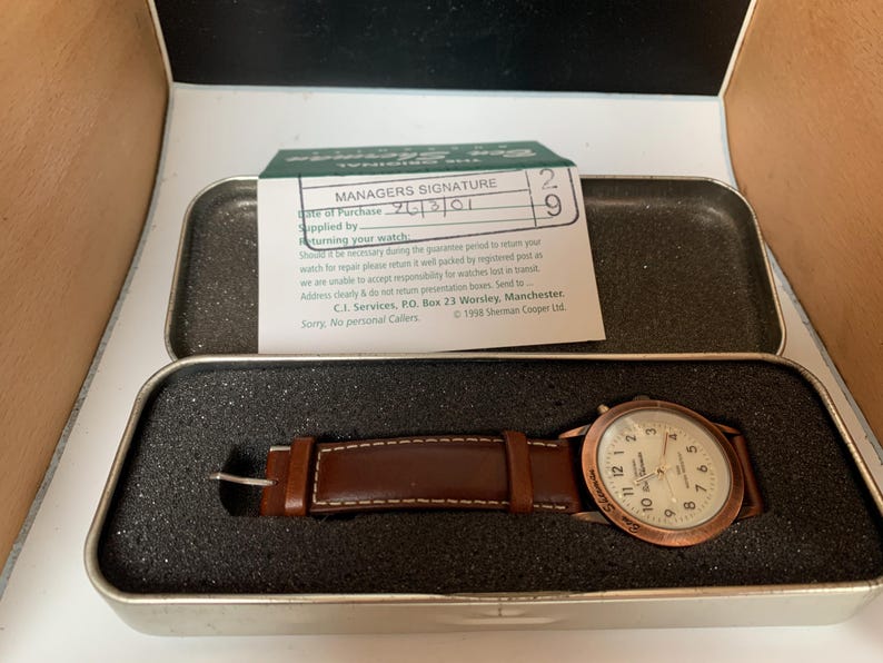 K&ouml;nnte beinhalten: Eine Vintage-Armbanduhr mit braunem Lederarmband und kupferfarbenem Geh&auml;use, pr&auml;sentiert in einer Metalldose. Das Zifferblatt der Uhr ist wei&szlig; mit schwarzen Ziffern. Eine kleine Karte mit Text ist sichtbar.