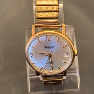May include: A vintage gold-toned Ramona wristwatch with a silver face, featuring the date 13. The watch has a gold expanding band. The watch face displays the text "RAMONA 25 JEWELS INCABLOC AUTOMATIC".