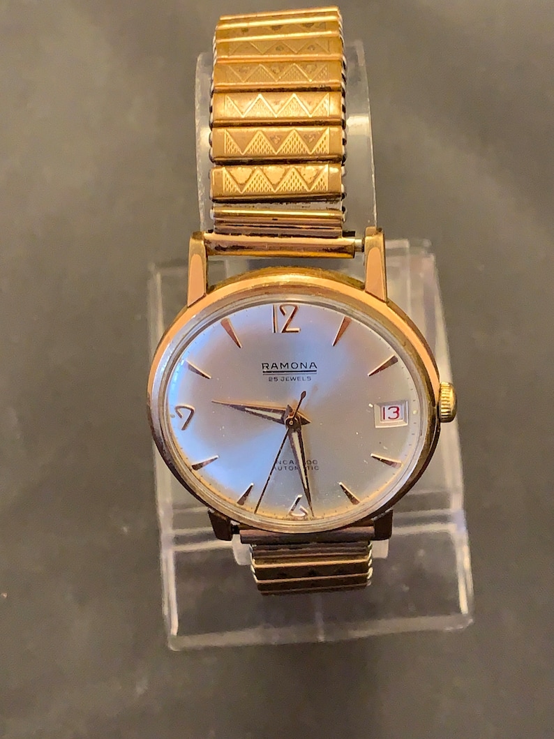 May include: A vintage gold-toned Ramona wristwatch with a silver face, featuring the date 13. The watch has a gold expanding band. The watch face displays the text "RAMONA 25 JEWELS INCABLOC AUTOMATIC".