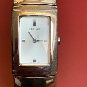 Könnte beinhalten: Eine silberne Pulsar-Armbanduhr mit einem rechteckigen weißen Zifferblatt und schwarzen Zeigern. Die Uhr hat ein silbernes Armband mit einem rechteckigen Design.
