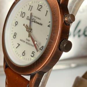 K&ouml;nnte beinhalten: Eine Ben Sherman Uhr mit kupferfarbenem Geh&auml;use und braunem Lederarmband. Das Zifferblatt ist cremefarben mit schwarzen Ziffern und Zeigern. Der Text "Ben Sherman" und "50M Water Resistant" sind sichtbar.