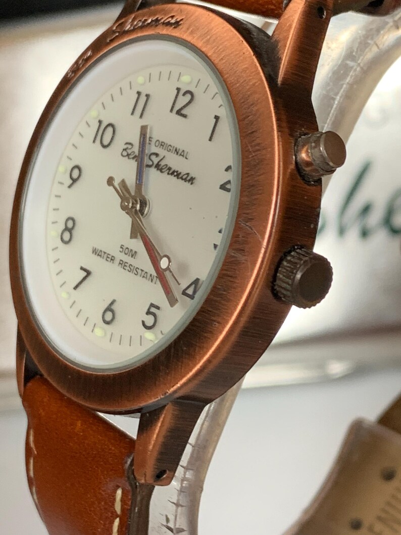 K&ouml;nnte beinhalten: Eine Ben Sherman Uhr mit kupferfarbenem Geh&auml;use und braunem Lederarmband. Das Zifferblatt ist cremefarben mit schwarzen Ziffern und Zeigern. Der Text "Ben Sherman" und "50M Water Resistant" sind sichtbar.