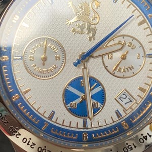 Puede incluir: Primer plano de un reloj de pulsera plateado con esfera blanca y detalles azules y dorados. El reloj presenta m&uacute;ltiples subesferas, incluida una con el texto "SCOTLAND FOREVER". Un emblema de le&oacute;n dorado en la parte superior. La fecha se muestra a las 3 en punto.