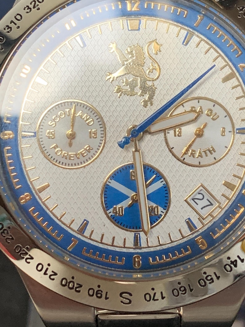 Puede incluir: Primer plano de un reloj de pulsera plateado con esfera blanca y detalles azules y dorados. El reloj presenta m&uacute;ltiples subesferas, incluida una con el texto "SCOTLAND FOREVER". Un emblema de le&oacute;n dorado en la parte superior. La fecha se muestra a las 3 en punto.