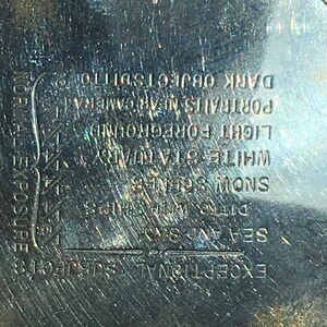 Op de afbeelding: Close-up van een zilverkleurig metalen object met ge&euml;tste tekst. De tekst bevat zinnen als "Uitzonderlijke onderwerpen", "Zee en lucht" en "Sneeuwsc&egrave;nes". Het object heeft een cirkelvorm met een getextureerde rand.