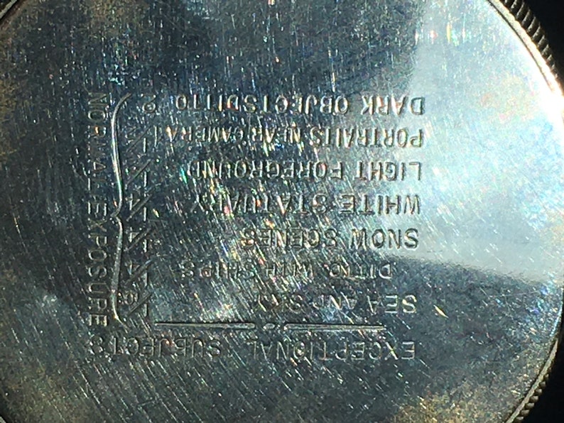 Op de afbeelding: Close-up van een zilverkleurig metalen object met ge&euml;tste tekst. De tekst bevat zinnen als "Uitzonderlijke onderwerpen", "Zee en lucht" en "Sneeuwsc&egrave;nes". Het object heeft een cirkelvorm met een getextureerde rand.