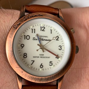K&ouml;nnte beinhalten: Ben Sherman Uhr mit braunem Lederarmband und kupferfarbenem Geh&auml;use. Das wei&szlig;e Zifferblatt hat schwarze Ziffern und Zeiger. Auf dem Zifferblatt steht "Ben Sherman" und "Water Resistant".