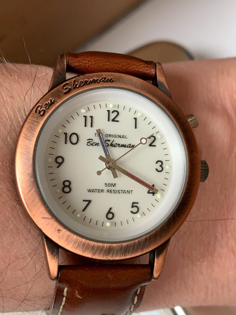 K&ouml;nnte beinhalten: Ben Sherman Uhr mit braunem Lederarmband und kupferfarbenem Geh&auml;use. Das wei&szlig;e Zifferblatt hat schwarze Ziffern und Zeiger. Auf dem Zifferblatt steht "Ben Sherman" und "Water Resistant".
