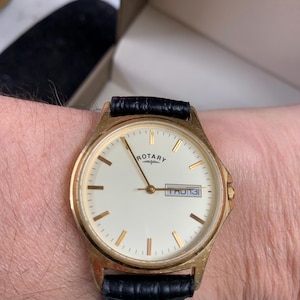 Puede incluir: Un reloj Rotary dorado con esfera blanca y correa de cuero negro. El reloj muestra el día y la fecha, con la palabra "ROTARY" impresa encima de las manecillas. El reloj está en una muñeca.