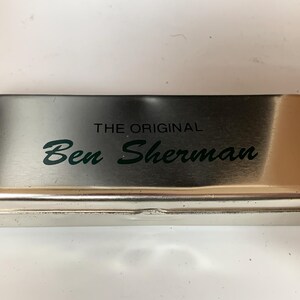 K&ouml;nnte beinhalten: Ein rechteckiges, silberfarbenes Federm&auml;ppchen aus Metall mit abgerundeten Kanten. Der Text "THE ORIGINAL" steht &uuml;ber dem Namen "Ben Sherman" in gr&uuml;ner, kursiver Schrift. Das Etui hat eine leicht reflektierende Oberfl&auml;che.