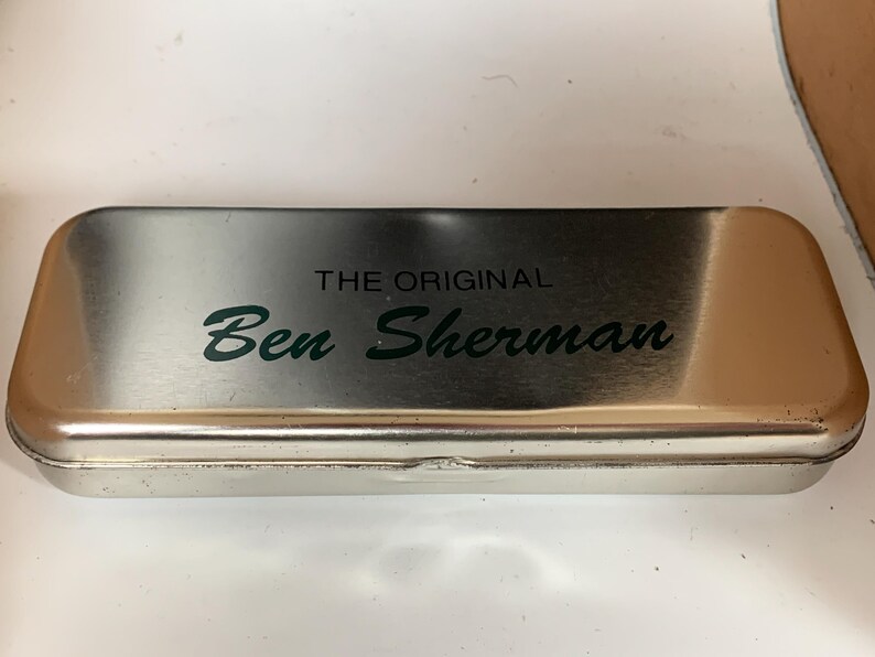 K&ouml;nnte beinhalten: Ein rechteckiges, silberfarbenes Federm&auml;ppchen aus Metall mit abgerundeten Kanten. Der Text "THE ORIGINAL" steht &uuml;ber dem Namen "Ben Sherman" in gr&uuml;ner, kursiver Schrift. Das Etui hat eine leicht reflektierende Oberfl&auml;che.